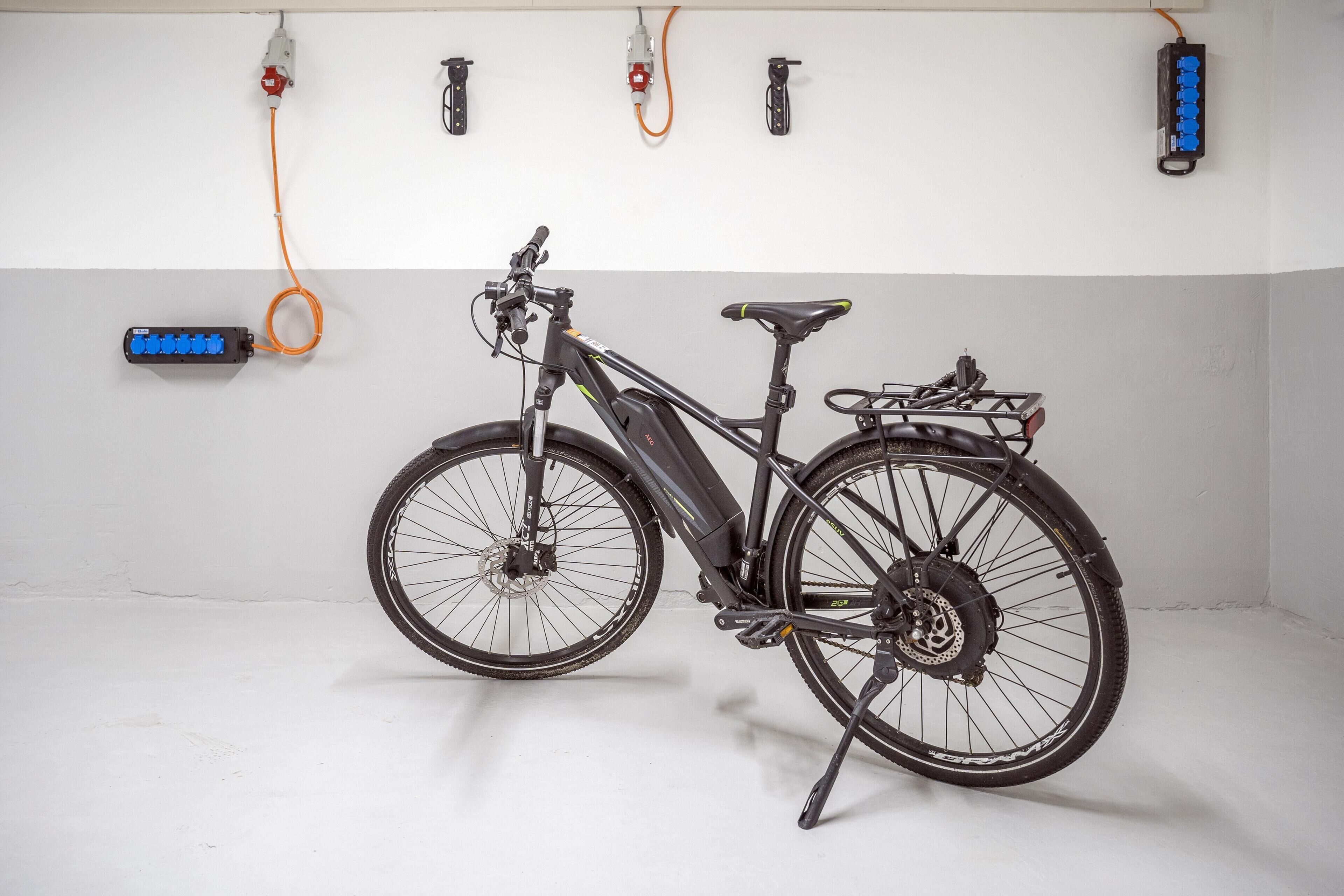 A stationary electric bicycle with a rear rack, connected to a wall-mounted charging station.