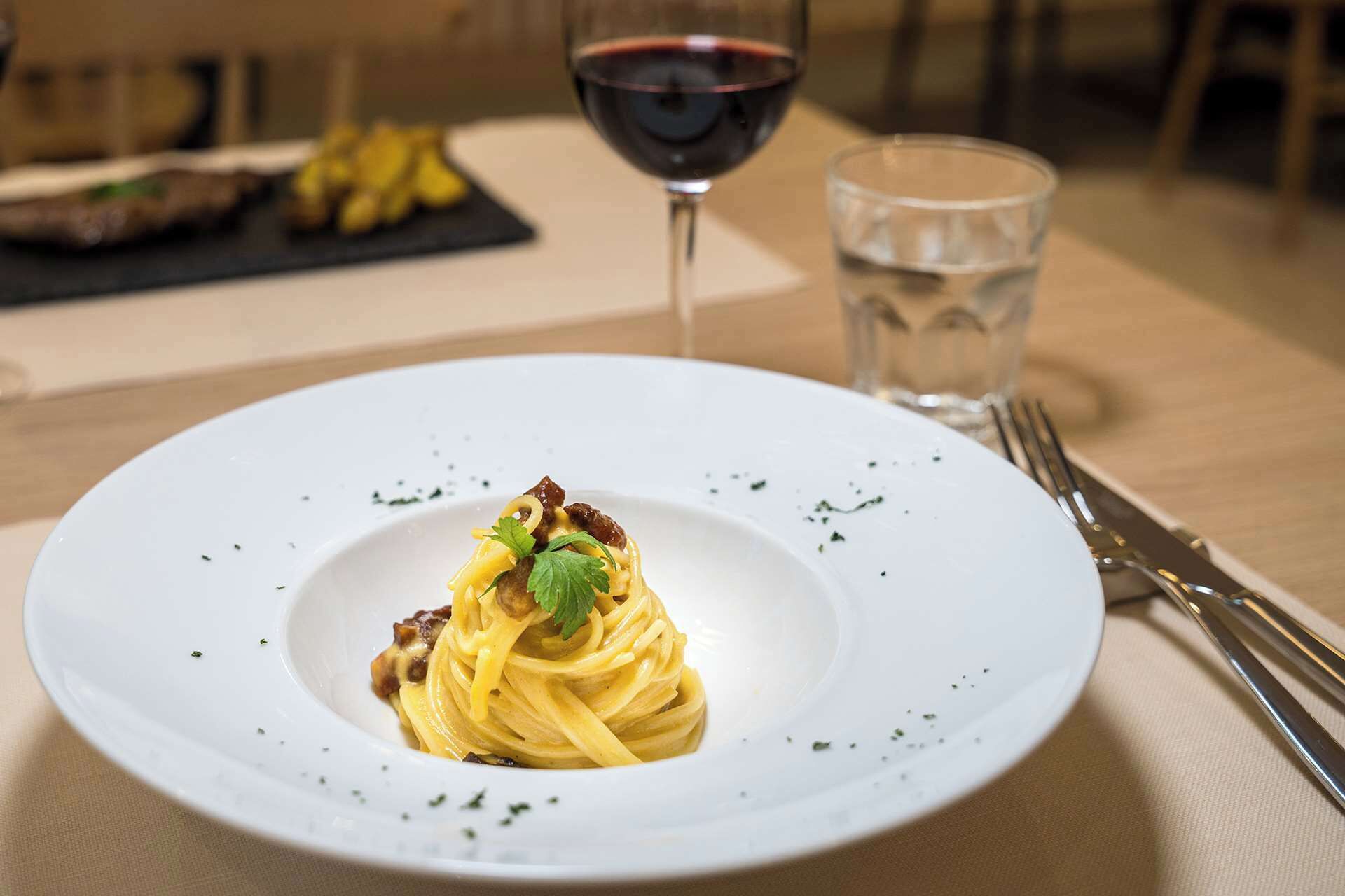 Un plato de pasta con salsa y decoración, una copa de vino tinto y un vaso de agua sobre una mesa.