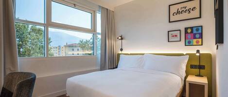 A hotel room with a large bed, a chair, a bedside table, a lamp, and a view of buildings through the window.