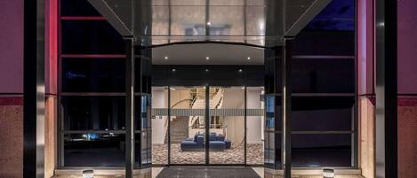 Una entrada de hotel moderna con fachada de vidrio, una escalera central y un área de descanso con sofá y sillones.