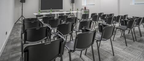 A conference room with rows of black chairs, a large flat-screen TV, and a white table with bottles.