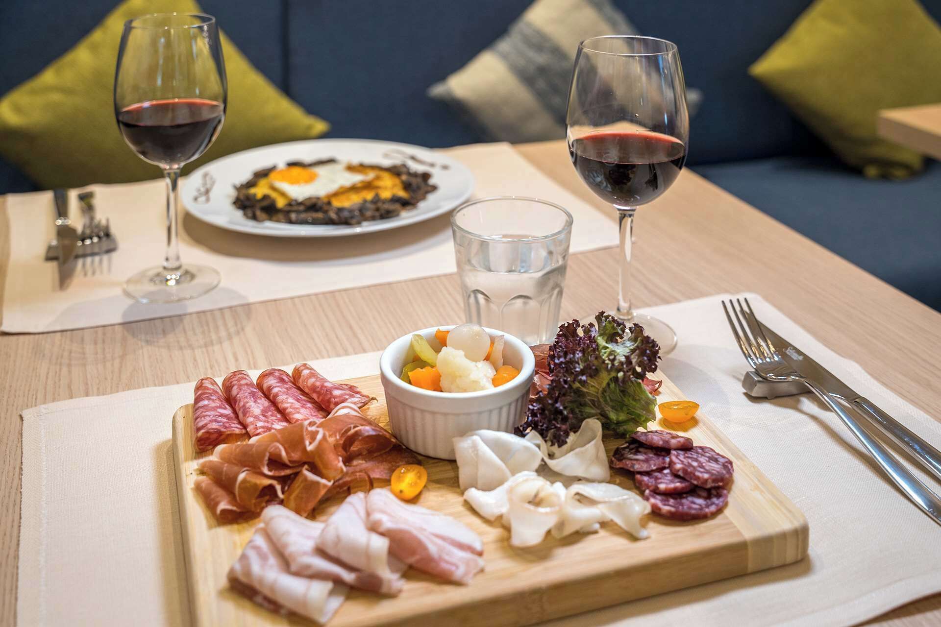 A breakfast setting with a variety of meats, a bowl of fruit, and two glasses of red wine on a wooden table.