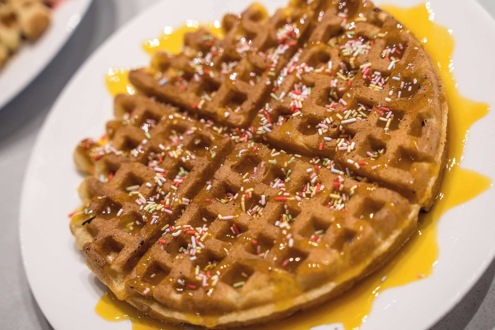 A close-up of a waffle with sprinkles and syrup.