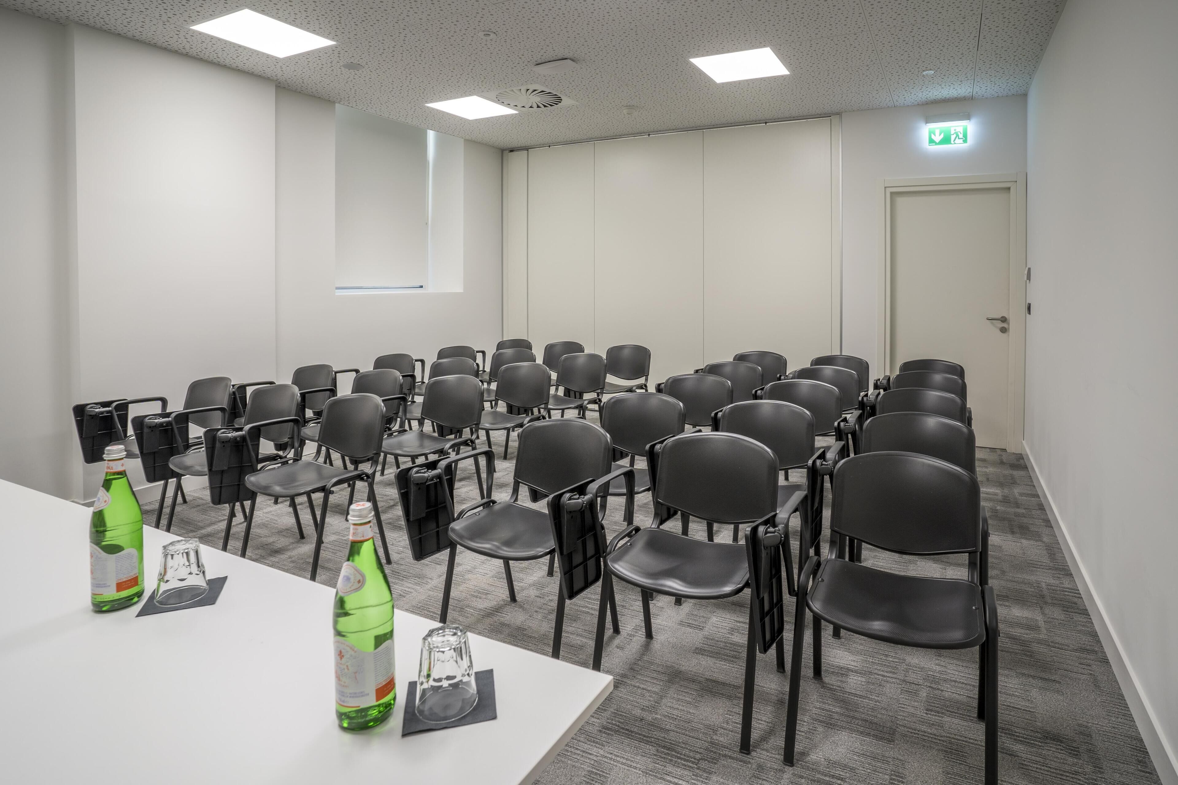 Une salle de conférence avec des rangées de chaises, une table blanche sur laquelle sont posées deux bouteilles d’eau, et une fenêtre.