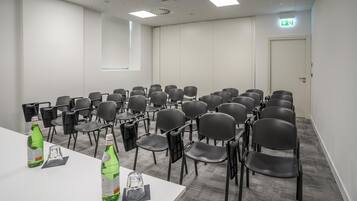 Une salle de conférence avec des rangées de chaises, une table blanche sur laquelle sont posées deux bouteilles d’eau, et une fenêtre.