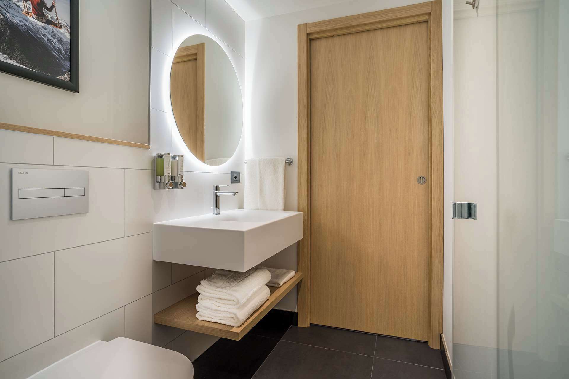 A modern bathroom with a white vanity, a large mirror, and a wooden door.
