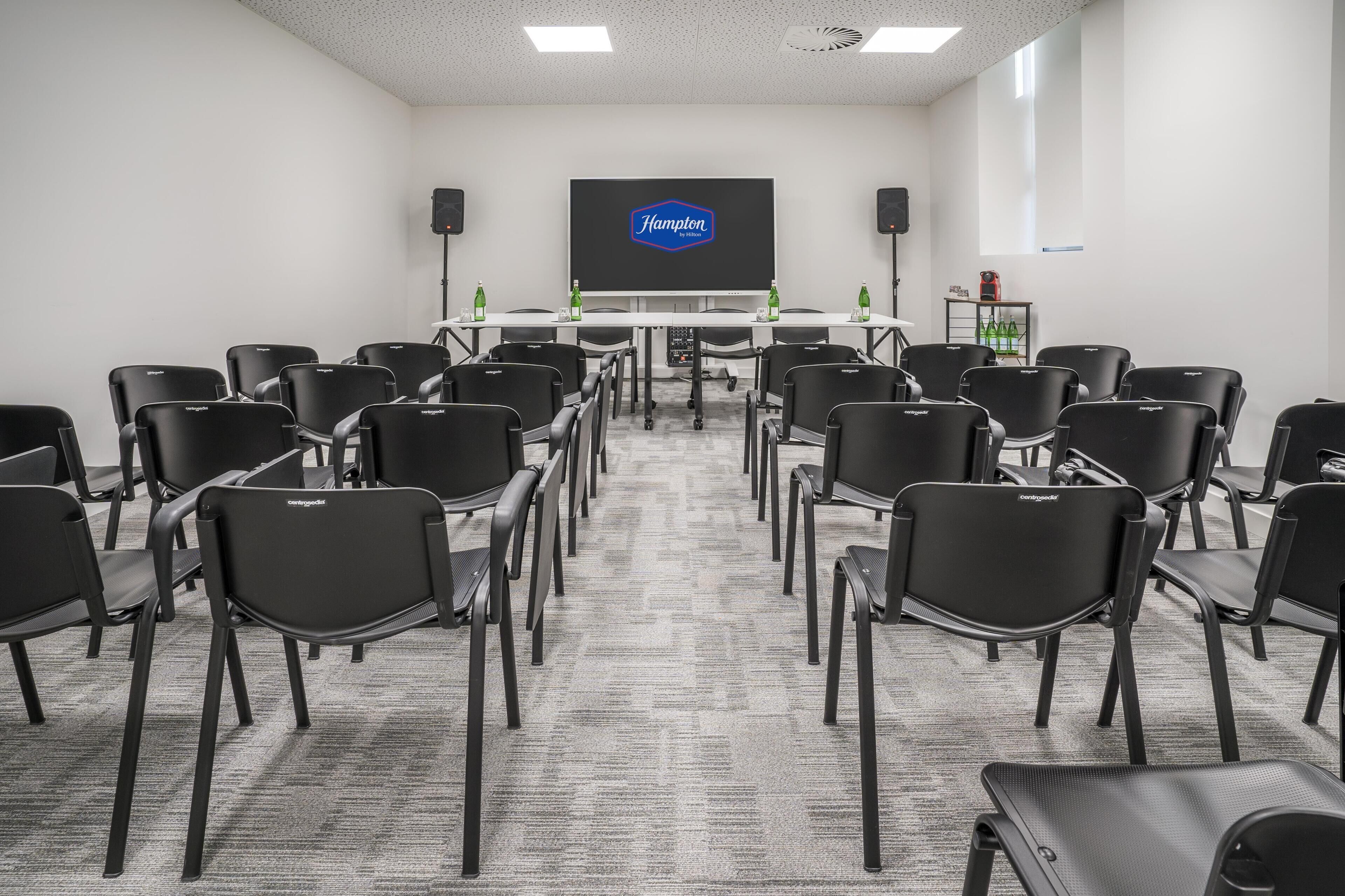 A conference room with black chairs, a large screen displaying 'Hampton,' and a carpeted floor.