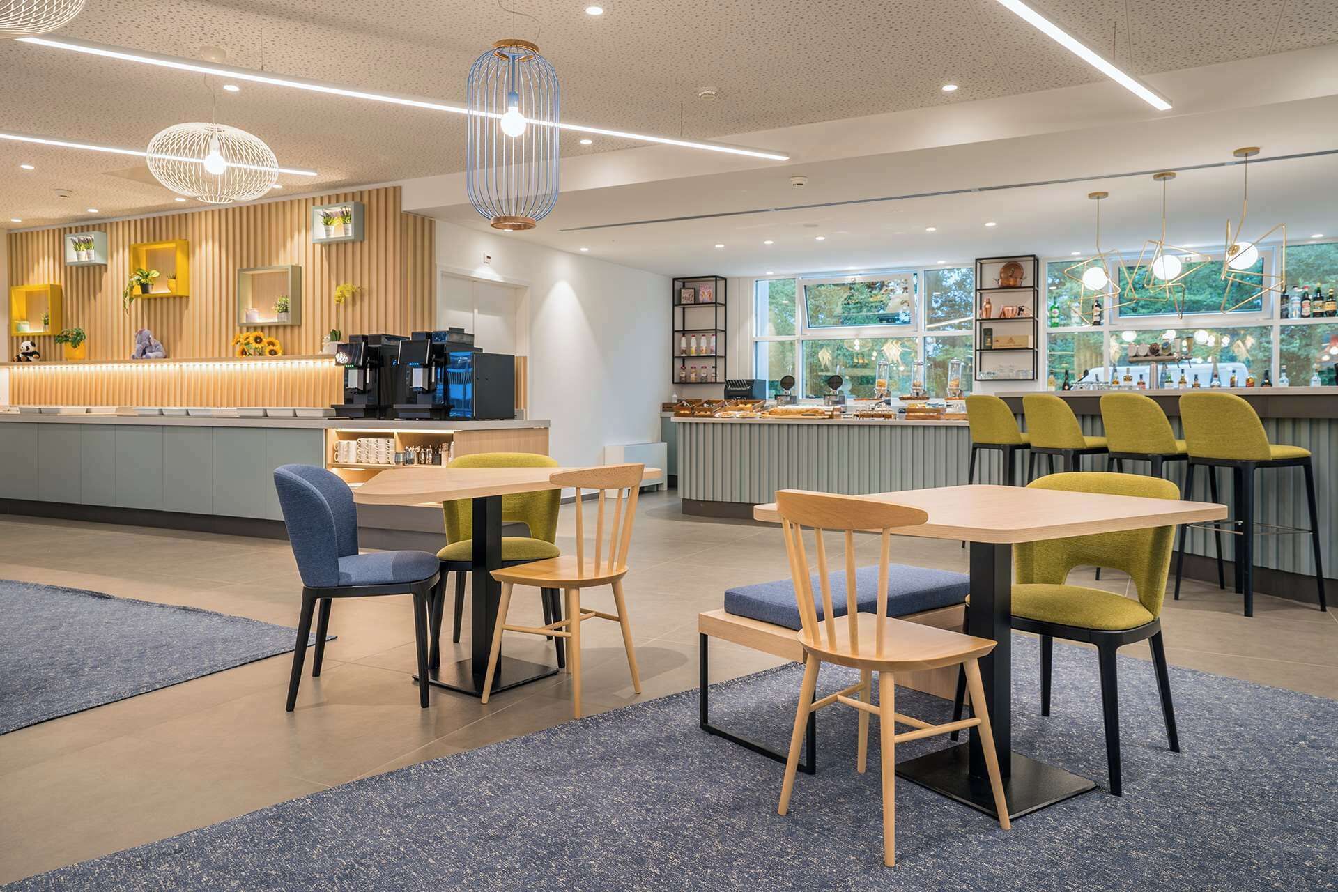 Un café moderno con mesas y sillas de madera, una barra de café y ventanas amplias que ofrecen vistas a zonas verdes.