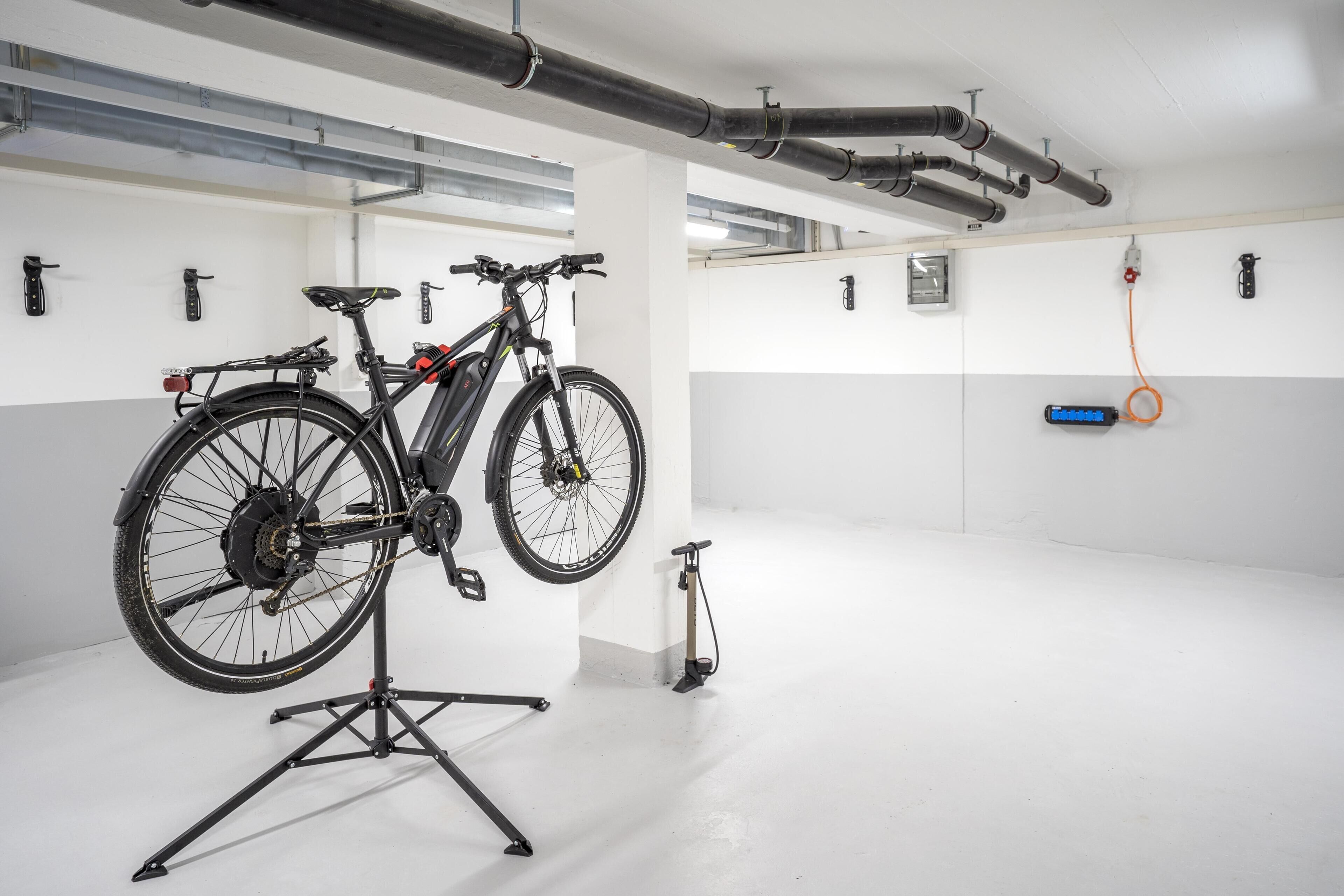 Una bicicletta fissata a un supporto in un ambiente interno pulito, con pareti bianche, tubature a vista e un dispositivo blu al muro.