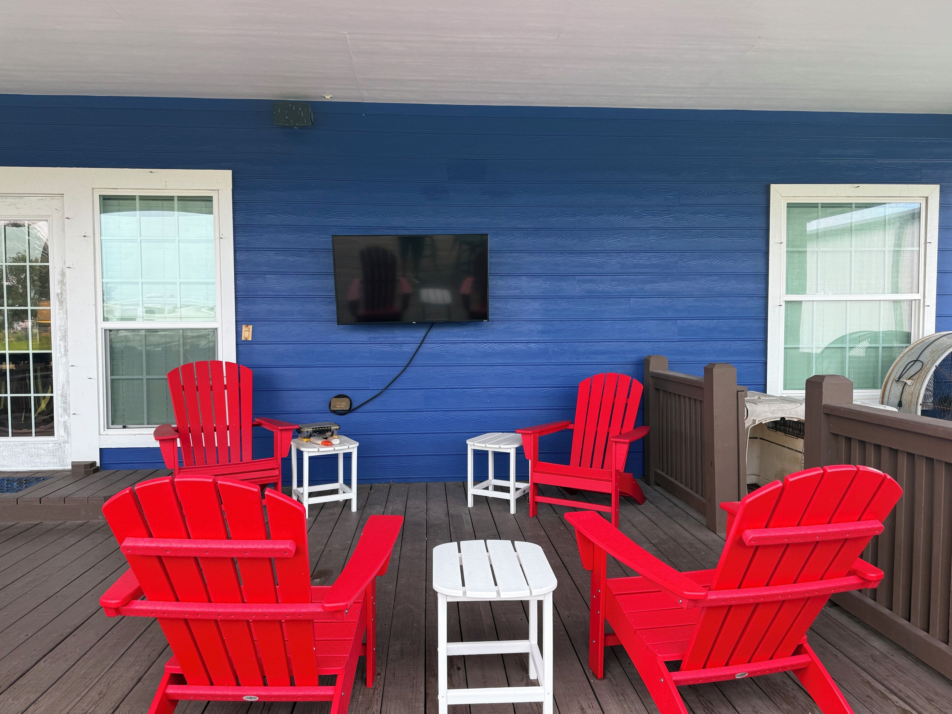 Un espacio de estar al aire libre con techo, sillas Adirondack rojas y una mesa blanca, un televisor montado en la pared y una terraza de madera.