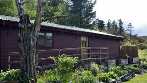 A rustic cabin with a wooden deck, surrounded by a well-maintained garden and a large tree.