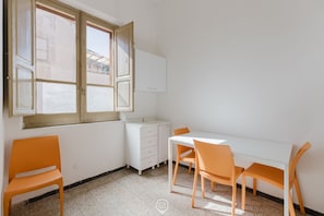 Una habitación pequeña y luminosa con un escritorio blanco, dos sillas naranjas, un armario blanco y una ventana con contraventanas abiertas.