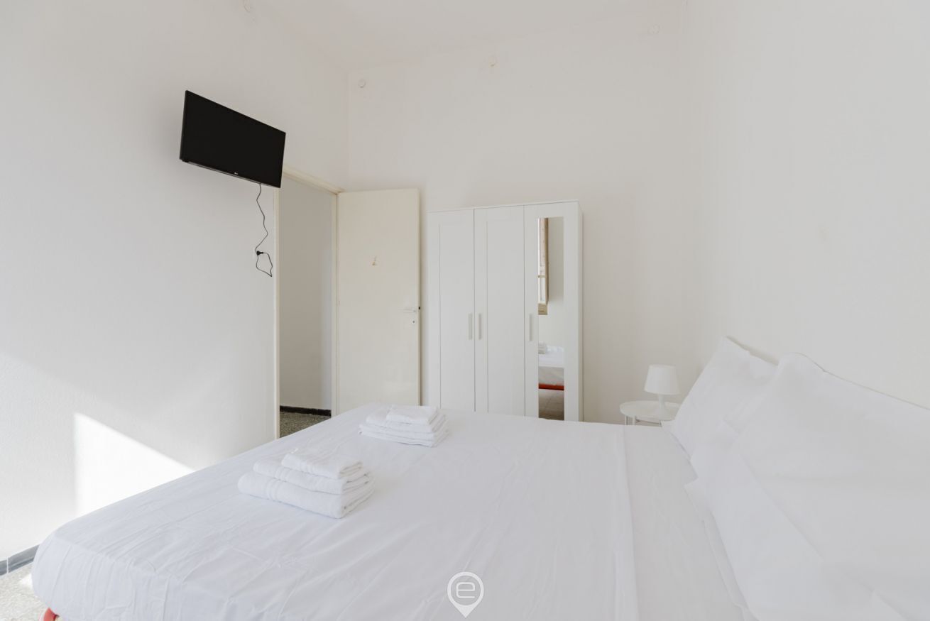 A hotel room with a bed, a TV mounted on the wall, a wardrobe, and a small table with a lamp.