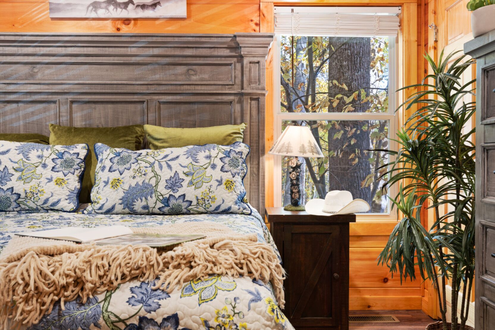 Un dormitorio con cabecera de madera, ropa de cama floral, una lámpara de noche y ventana con vistas a árboles.