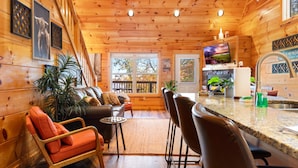Interior acogedor de una cabaña con techo de madera, una sala de estar con sofá y sillones, una cocina con barra y vistas al exterior a través de amplios ventanales.