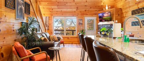 A cozy cabin interior with a wooden ceiling, a living area with a sofa and chairs, a kitchen with a bar, and a view of the outdoors through large windows.
