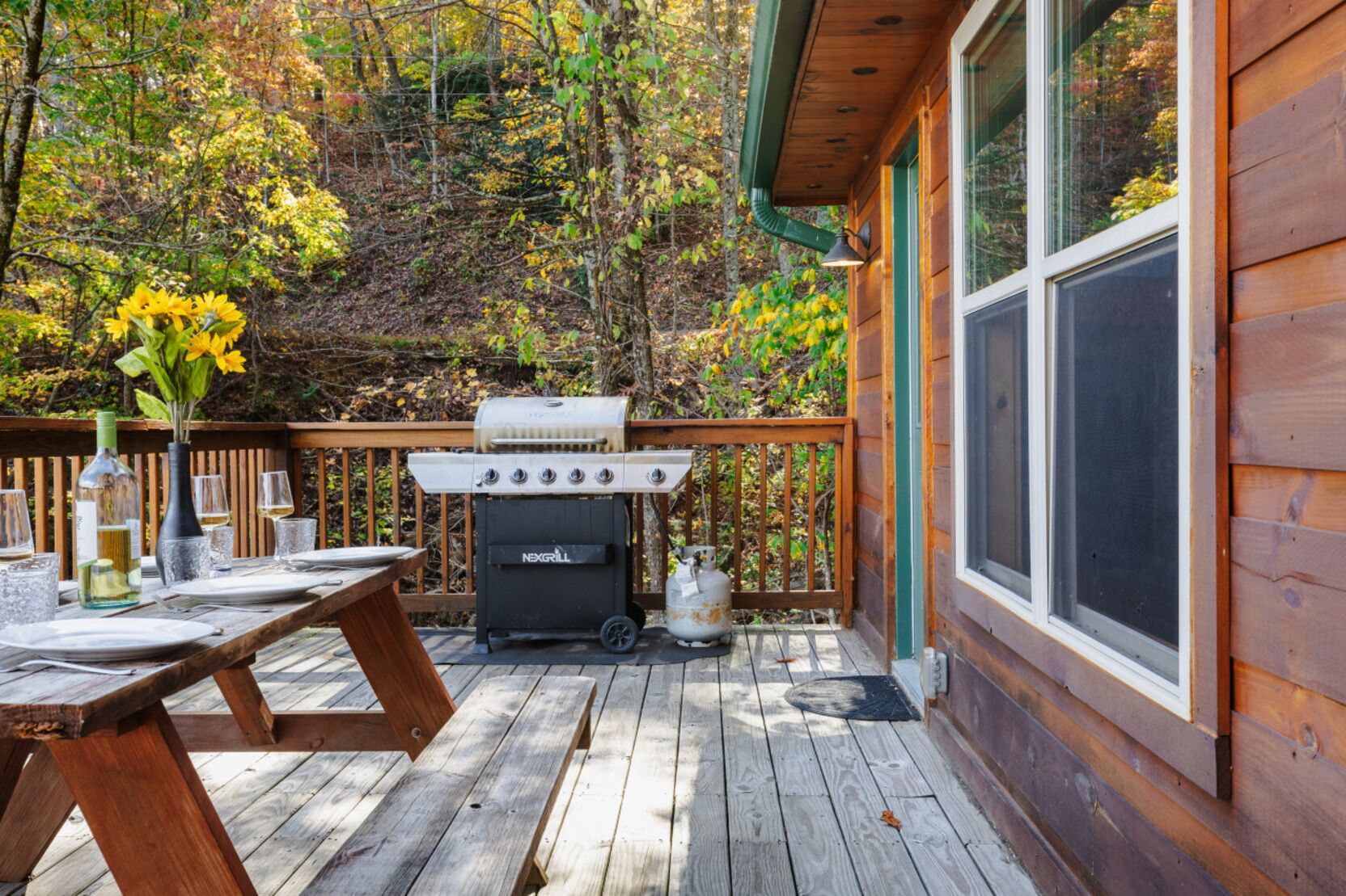 A wooden deck with a grill, a table set for dining, and a view of a forested area.