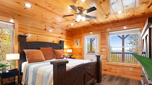 Dormitorio de cabaña de madera con cama, mesitas de noche, ventilador de techo y ventanas amplias que ofrecen vistas al exterior.