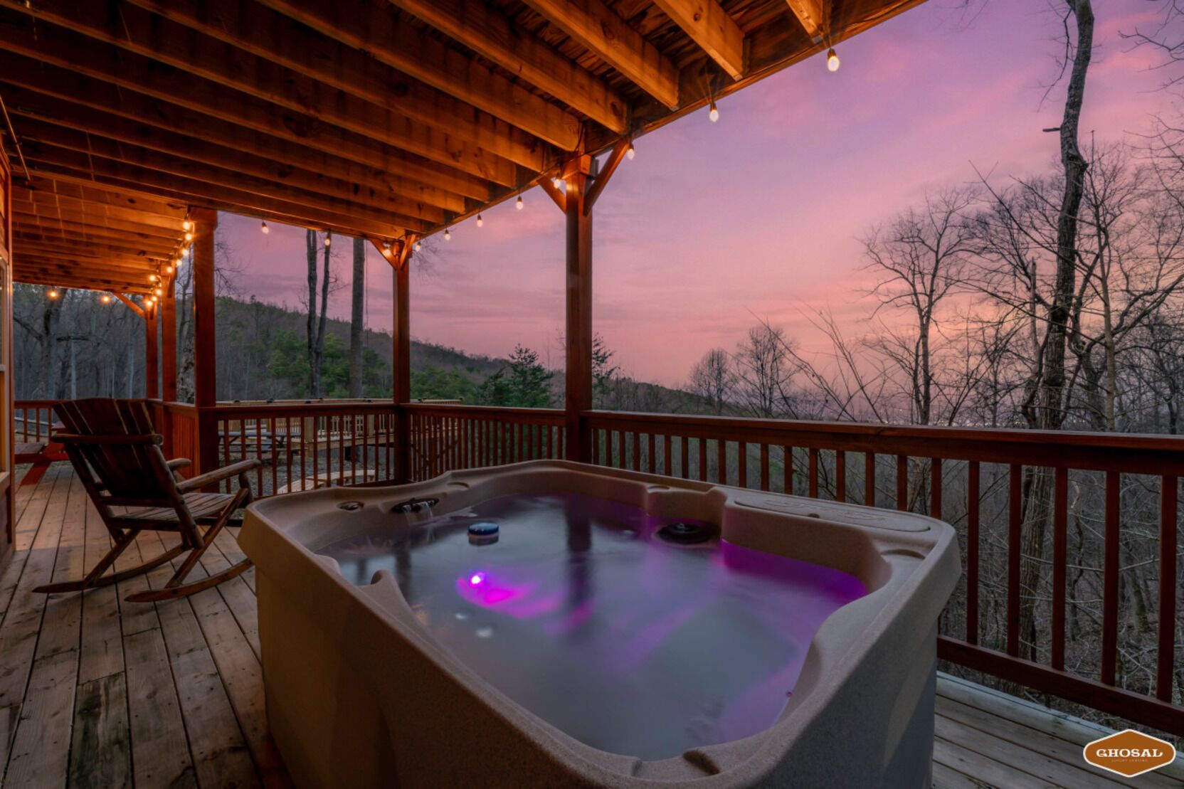 Eine Holzterrasse mit Whirlpool und Schaukelstuhl, mit Blick auf eine bewaldete Landschaft bei Sonnenuntergang.