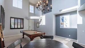 A spacious room with a pool table, a chandelier, and a large window with blinds. - Elegant desert getaway. 4 bedroom 2.5 bath. Sleeps 8. Heated pool. Outdoor BBQ. (Oro Valley)