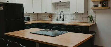 A modern kitchen with white cabinets, a black refrigerator, a microwave, a sink with a black faucet, and a wooden island with chairs.