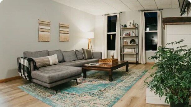Ein Wohnzimmer mit grauem Sofa, einem Holztisch, einem gemusterten Teppich, einer Stehlampe und einer Topfpflanze.