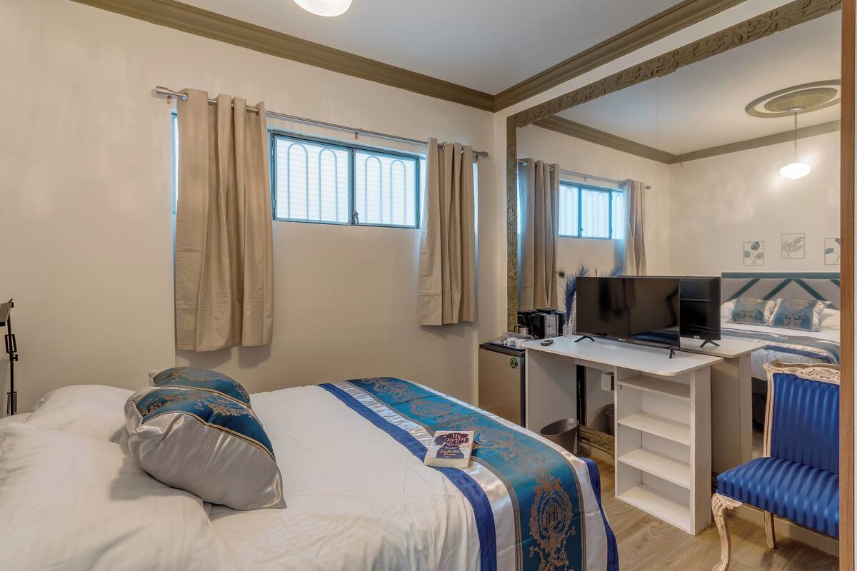 A hotel room with a bed, a desk with a TV, a chair, and a small desk lamp.