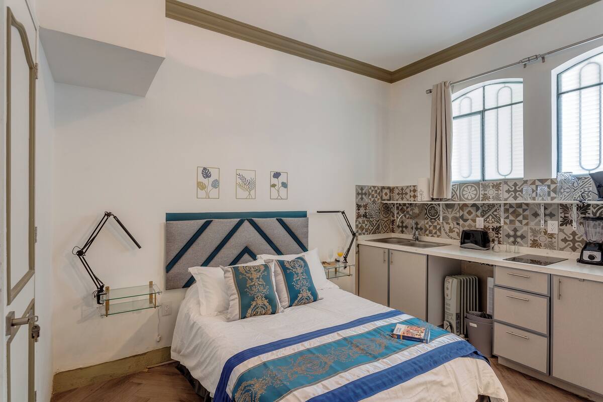 A hotel room with a bed, bedside table, lamp, and a view of the kitchen area.