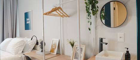 Una habitación de hotel moderna con una cama, un escritorio con una silla, un espejo redondo, un lavamanos pequeño y una planta.