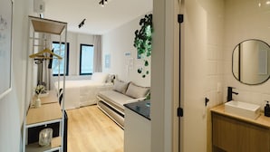Una habitación de hotel moderna con cama, sofá, escritorio con silla, pequeña cocina y baño con lavamanos y espejo.
