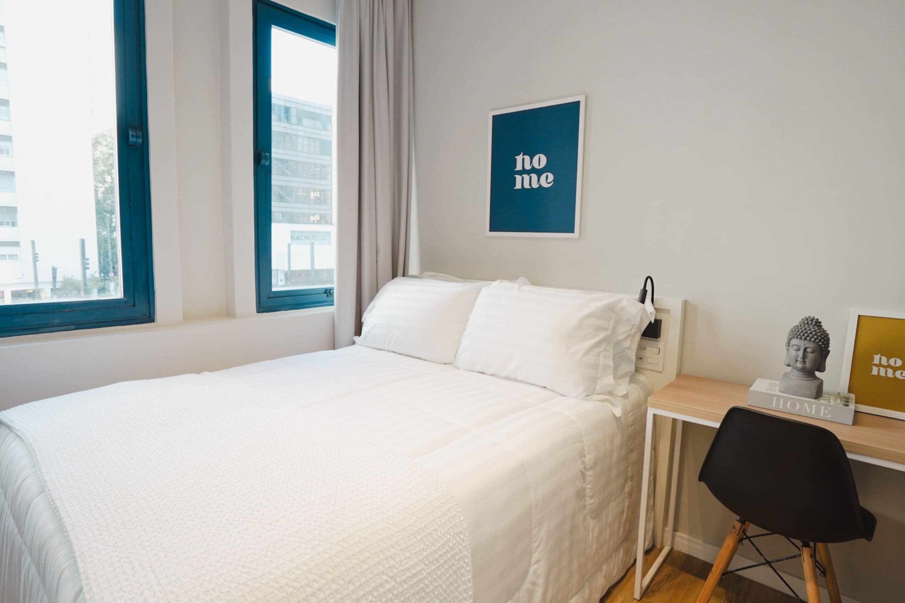 A neatly made bed with white bedding, a small desk with a chair, and a window with a view of buildings outside.