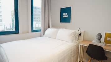 A neatly made bed with white bedding, a small desk with a chair, and a window with a view of buildings outside.