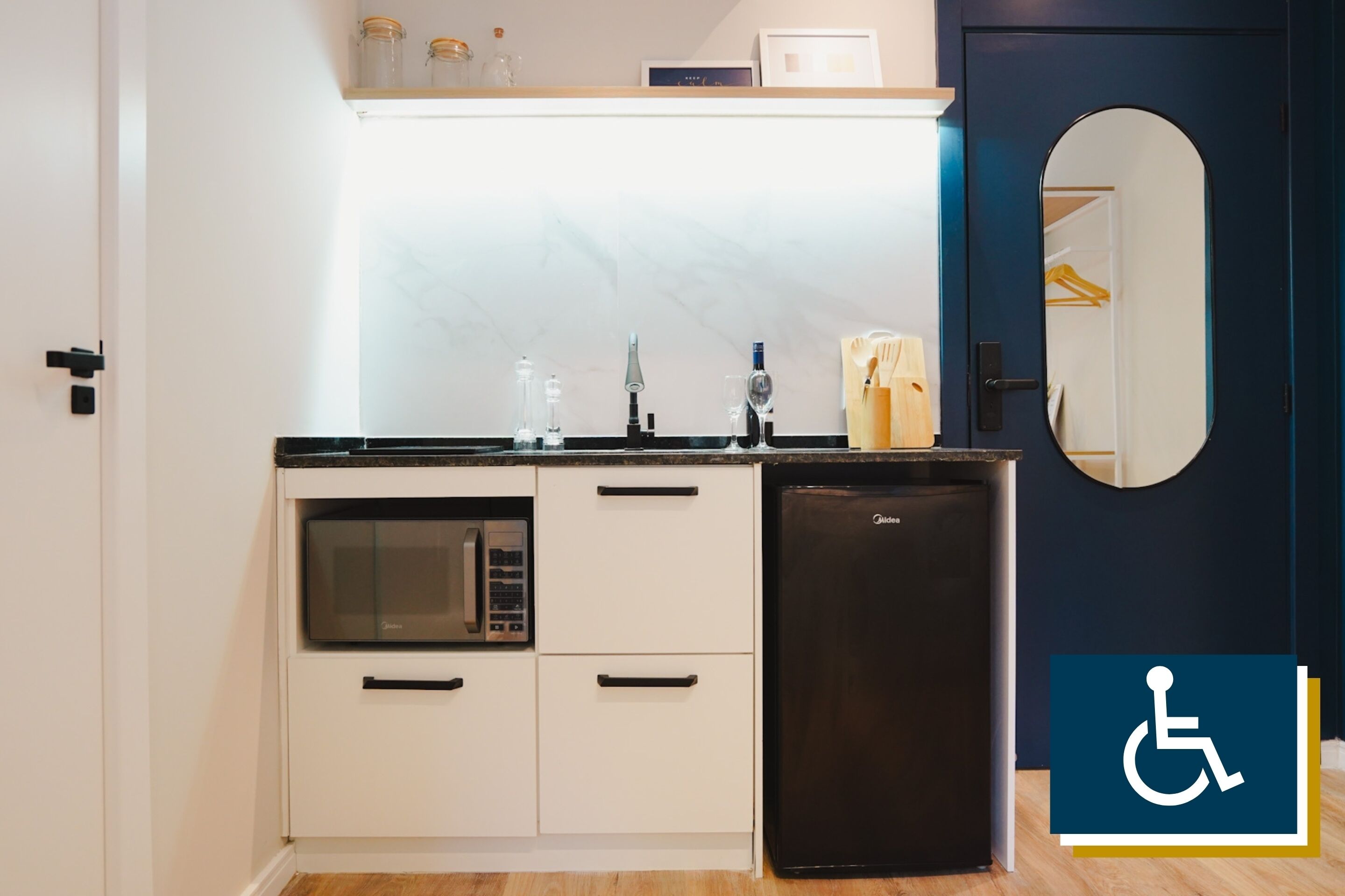 A compact kitchen with a microwave, oven, and refrigerator. There is a shelf above the sink with jars and a small clock. A mirror hangs on the wall to the right.