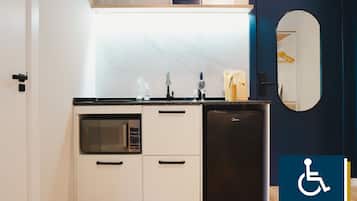 Cocina compacta con microondas, horno y refrigerador. Hay una repisa sobre el lavamanos con frascos y un pequeño reloj. Un espejo cuelga en la pared a la derecha.