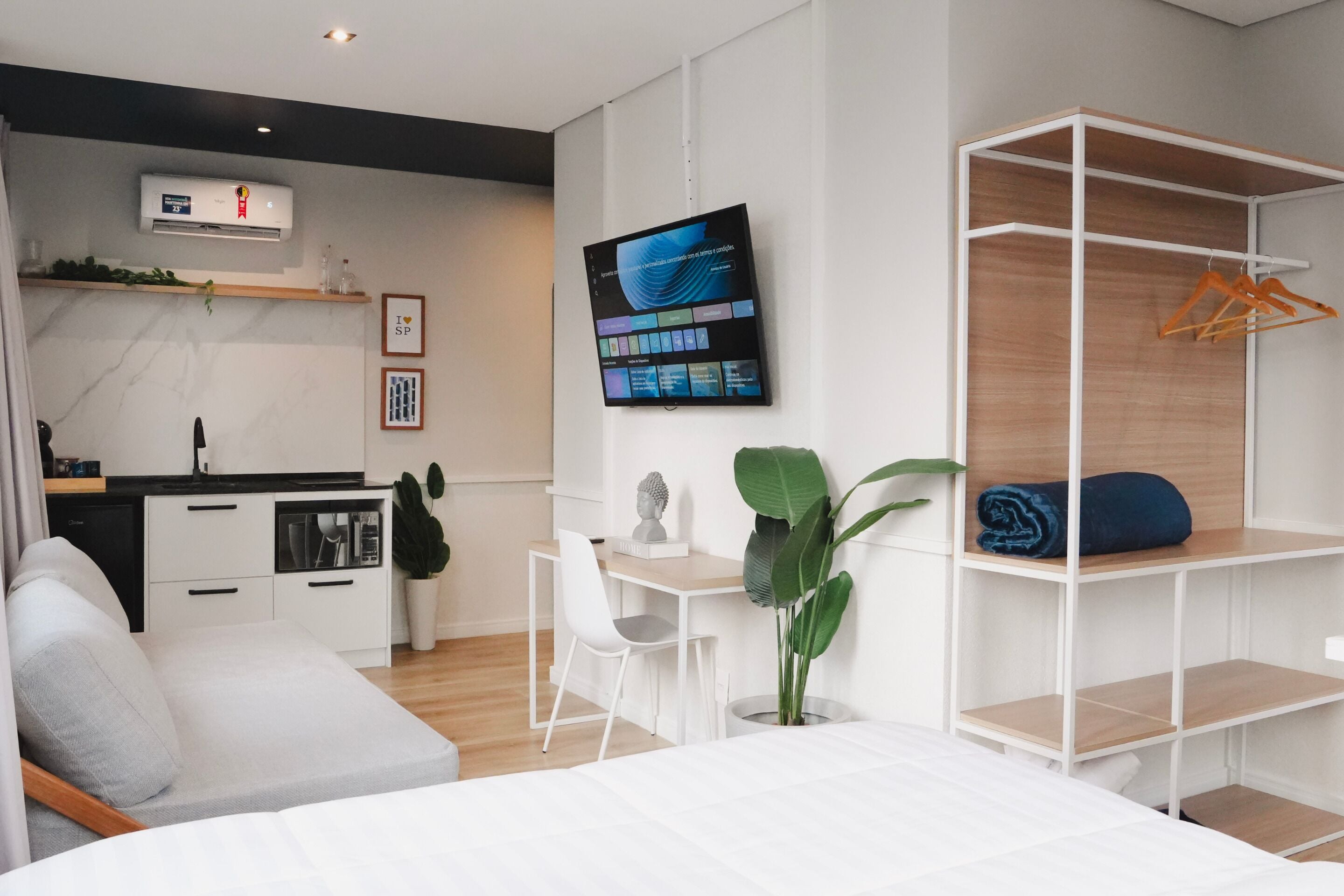 A modern hotel room with a bed, a desk, a chair, a TV, a wardrobe, and a small kitchen area.