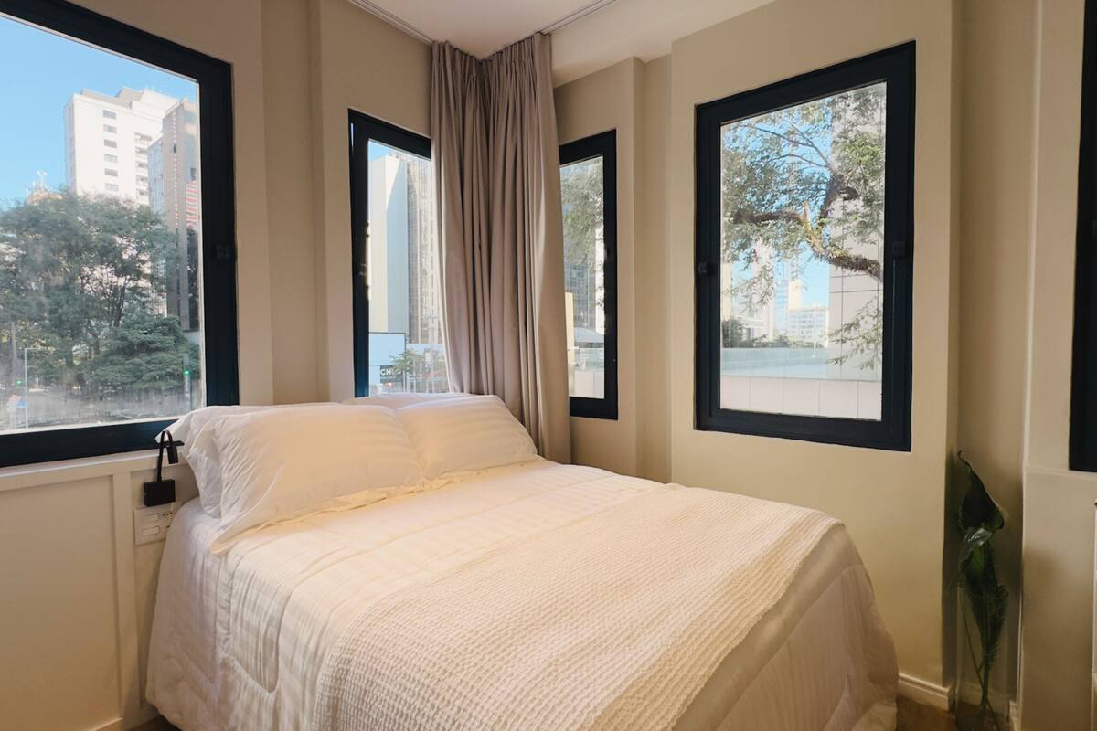 Ein Schlafzimmer mit Bett, großen Fenstern und Blick auf Gebäude und Bäume.