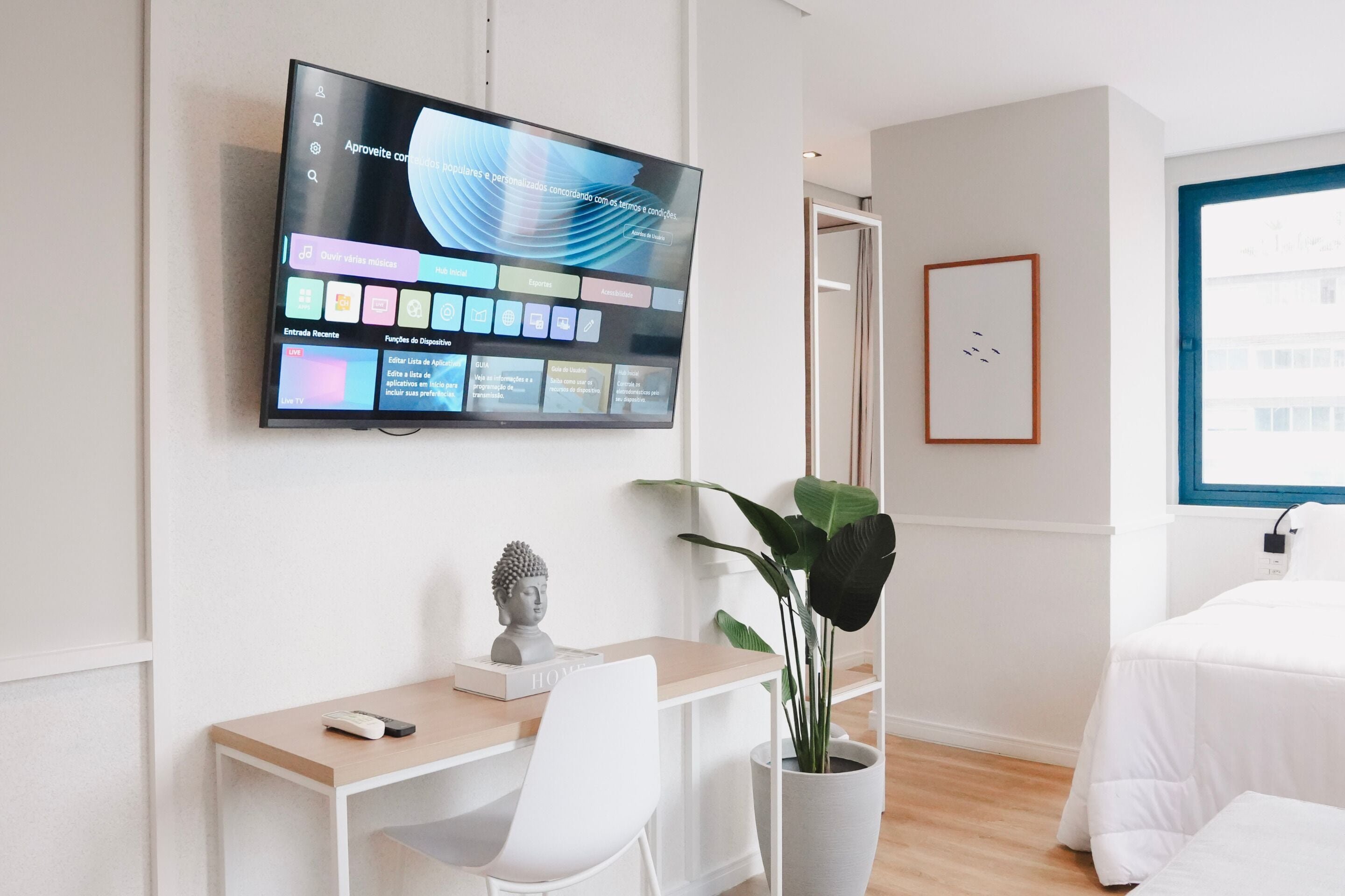Moderni hotellihuone, jossa on seinälle asennettu taulutelevisio, työpöytä tuolilla, ruukkukasvi ja sänky.