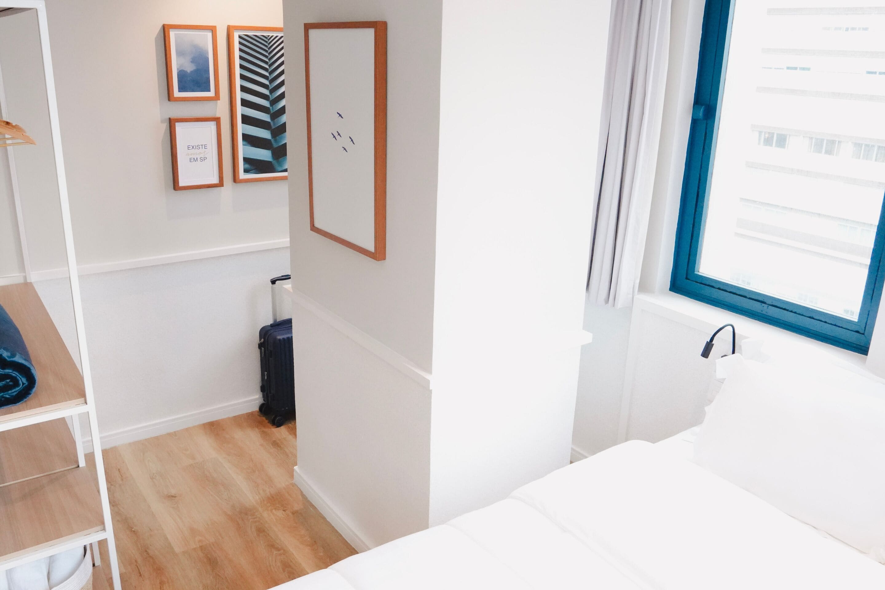 A hotel room with a bed, a desk, a wardrobe, a suitcase, and framed pictures on the wall.