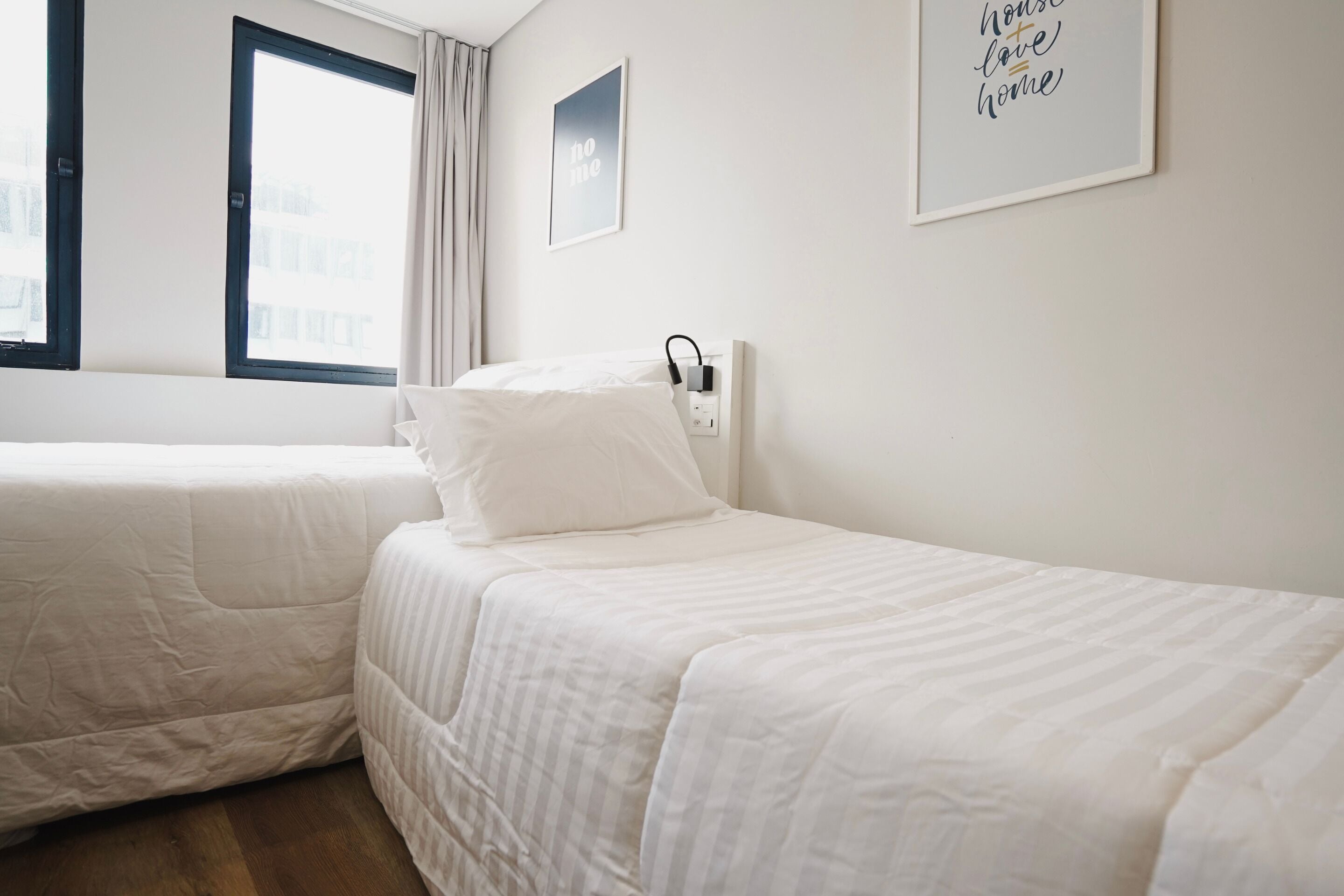 Una habitación de hotel con dos camas individuales, una ventana con cortinas, un enchufe en la pared y una cita enmarcadas en la pared.