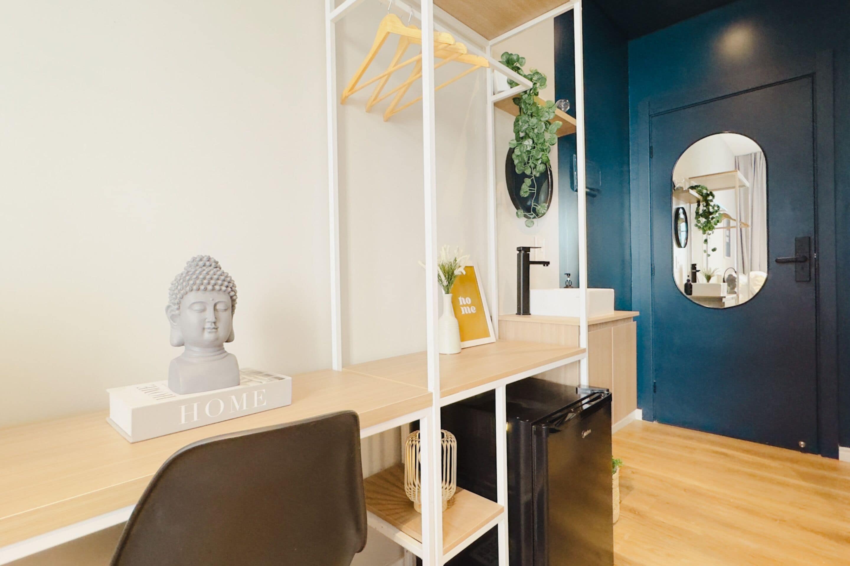 A modern room with a wooden desk, a black trash bin, a Buddha bust, a 'HOME' sign, a mirror, and a blue door.