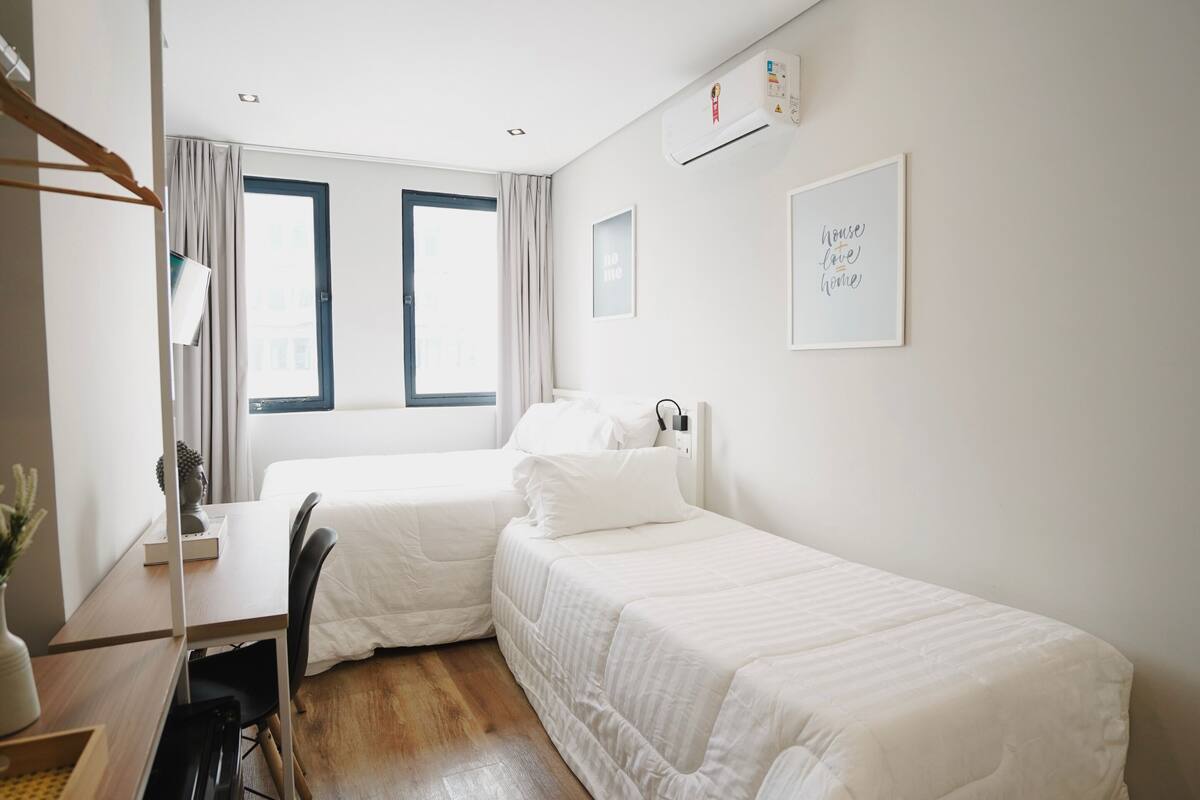 Ein Hotelzimmer mit zwei Einzelbetten, einem Schreibtisch mit Stuhl, einem kleinen Tisch mit Blumenvase und einem Fenster mit Vorhängen.