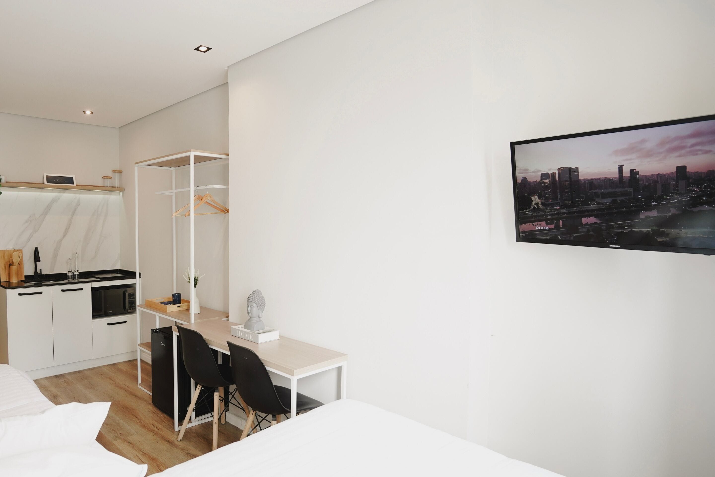 A modern hotel room with a bed, a desk, a chair, a wardrobe, a small kitchen area, and a cityscape view.
