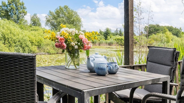 A wooden table with a vase of flowers and teapots, overlooking a pond with greenery and trees.
