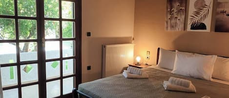 A bedroom with a large window, a bed with pillows, a bedside table with a lamp, and artwork on the wall.
