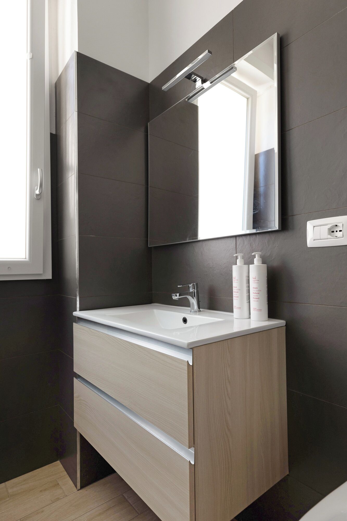 A modern bathroom with a white sink, dark tiled walls, and a large mirror.