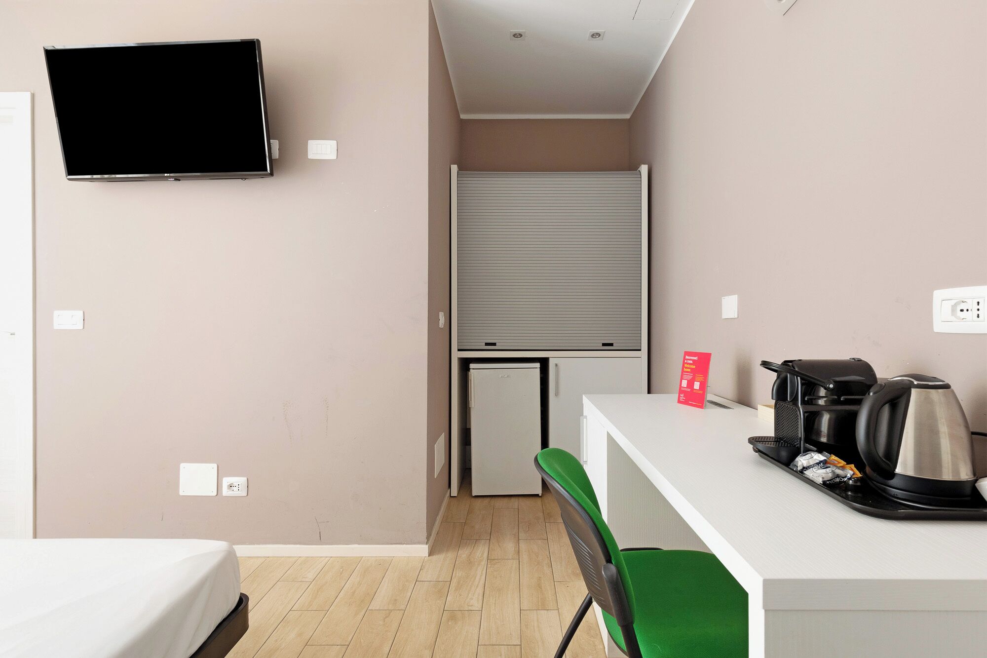 Ein kleines Hotelzimmer mit einem Bett, einem an der Wand befestigten Fernseher, einer Küchenzeile inklusive Kühlschrank, einer Kaffeemaschine und einem kleinen Tisch mit Stuhl.