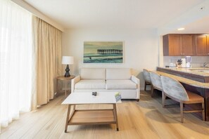 A modern hotel room with a sofa, a coffee table, a dining area with a counter and chairs, and a painting of a pier on the wall. - Pompano Beach | Couples Ocean Weekend | Pool + Beach (Pompano)
