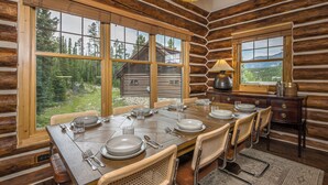 A log cabin dining room with a large wooden table set for six, large windows offering a view of the forest, and a chandelier hanging from the ceiling.