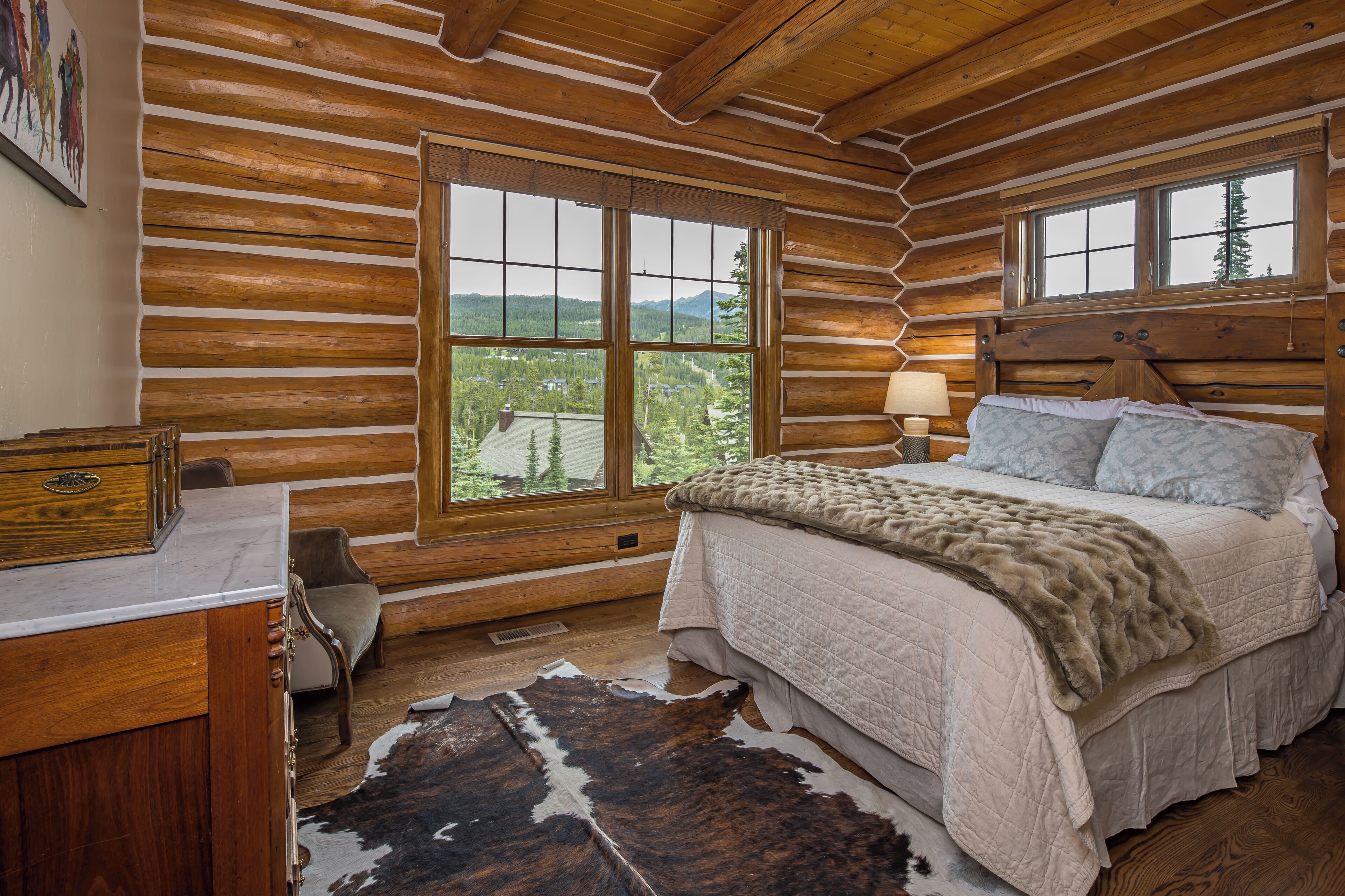A log cabin bedroom with a large bed, a wooden headboard, a window with a mountain view, and a cowhide rug.