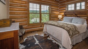 A log cabin bedroom with a large bed, a wooden headboard, a window with a mountain view, and a cowhide rug.